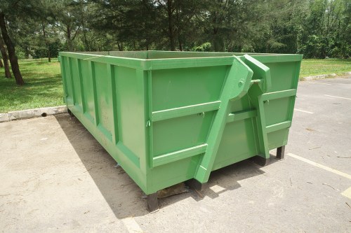 A large, rectangular green skip with metal sides and reinforced edges, positioned on a concrete driveway in an outdoor setting. The skip's surface has a smooth finish, and it is supported by a sturdy black base. The front features a hinged, metal handle mechanism to facilitate loading and unloading. Surrounding the skip, there is a neatly maintained grass verge and trees in the background, indicating a park or suburban area. The environment appears bright and clear, with natural daylight illuminating the scene. The skip is empty, ready for rubbish collection, aligning with services provided by Commercial Waste Bermondsey for waste removal in London. The overall scene emphasizes a professional and functional waste management setup consistent with local rubbish disposal needs.