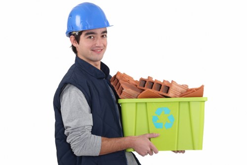 A young male waste management worker wearing a blue safety helmet and a navy blue vest over a grey long-sleeved shirt is smiling while holding a bright green recycling bin filled with red clay bricks. The bin features a blue recycling symbol on the side, indicating waste sorting or disposal. The background is plain white, emphasizing the individual and the bin. The scene suggests a professional rubbish removal service, possibly operating in Bermondsey or nearby London areas, focusing on construction debris or general waste collection as part of commercial waste management. The worker's casual yet professional attire, combined with the environment of the image, aligns with service providers like Commercial Waste Bermondsey handling local rubbish clearance tasks within the Bermondsey postcode district, offering environmentally conscious waste removal solutions for businesses or construction sites in the area.