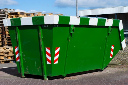 A large, green industrial rubbish skip positioned on a paved area outside, used for commercial waste removal in Bermondsey. The skip features white and green reflective safety markings along the top edges and sides, with a sturdy metal construction and a slightly tapered rectangular shape. It is situated near a chain-link fence and stacks of wooden pallets visible in the background, indicating an industrial or warehouse environment. The surface of the skip shows some minor scratches and paint finish typical of outdoor use. Behind the skip, there is a modern building with a corrugated metal roof and a lamp post, suggesting an industrial or commercial site in London’s Bermondsey postcode area. The scene is lit with natural daylight, highlighting the vibrant green colour and the textured metal surface of the waste container while reflecting subtle shadows cast on the pavement. This detailed description aligns with waste management services provided by Commercial Waste Bermondsey, emphasizing the size, material, markings, and setting for effective rubbish removal in the local area.