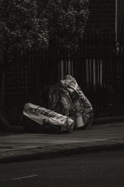 A narrow urban alleyway filled with a significant accumulation of waste materials, including large black and grey garbage bags and various cardboard boxes piled high at the far end of the space. In th