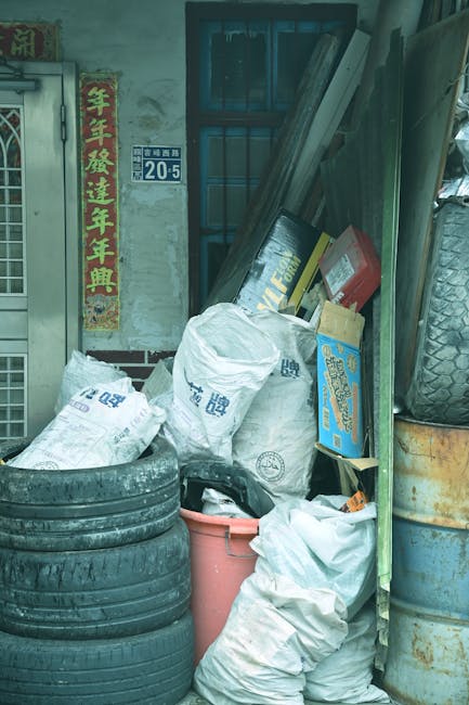 A red, corrugated metal dumpster with visible rust and weathering is positioned against a dark green wall and a light grey concrete wall within an urban alleyway or side street. Several white plastic