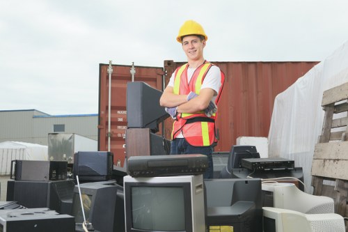 A young man wearing a yellow safety helmet and high-visibility vest stands outdoors at a waste disposal site, smiling with arms crossed amidst a collection of discarded electronic devices such as old computers, monitors, and printers. Behind him are shipping containers and stacks of wooden pallets, indicating an industrial or commercial environment typical of rubbish removal operations near Bermondsey. The electronic waste is piled on the ground, with various models and sizes of outdated technology clearly visible. The sky is slightly overcast, casting diffuse lighting over the scene, emphasizing the textures of the electronic components and the rugged outdoor setting. This image illustrates the process of rubbish clearance as handled by companies like Commercial Waste Bermondsey, highlighting their service in managing bulky and electronic waste pickups in the local area. The scene subtly reflects the importance of professional waste management solutions in urban environments focused on responsible disposal and recycling of electronic waste assets.
