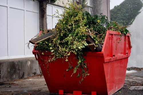 A large red plastic skip filled with assorted garden waste and debris, including leafy branches, plant clippings, and small wooden planks, situated outdoors on a paved surface next to a white brick wall with a large window, in an urban area potentially in Bermondsey. The skip's high sides are textured with visible ridges, and the waste materials overflow slightly at the top, with some greenery cascading over the edge. The background includes a concrete base and parts of a building, and the overall scene suggests a rubbish removal service managed by Commercial Waste Bermondsey for garden clearance or waste disposal in the local area.